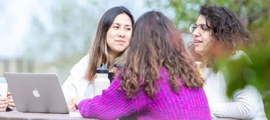 Three people pictured with a laptop are seated on a bench in a park and talking.