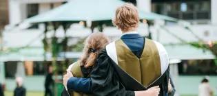 Two students wearing graduation gowns are hugging with their backs to the camera.