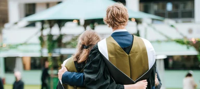 Two students wearing graduation gowns are hugging with their backs to the camera.