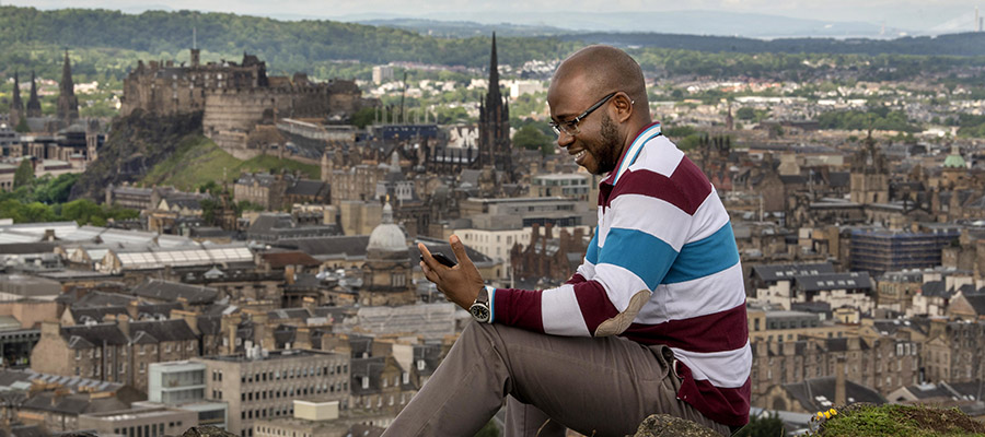 Student working on a laptop on the Salisbury Crags with Edinburgh skyline in the background.